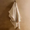 A beige waffle-weave towel hanging on a hook against a textured brown wall. Produktbild-Vorschau