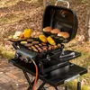 A portable gas grill with food cooking on it, set up outdoors on a wooden deck with autumn leaves on the ground. Produktbild-Vorschau