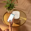 A person is holding a smartphone and interacting with a YABER T2 Plus LED projector on a round wooden table. A potted plant is also on the table. Produktbild-Vorschau