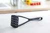 A black potato masher is placed on a white wooden table in a kitchen setting. In the background, a kitchen counter with some vegetables and a sink are visible. Náhľad obrázku produktu