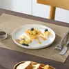 A dessert is presented on a white plate, set on a woven placemat on a wooden table. Cutlery is visible to the right of the plate. Produktbild-Vorschau