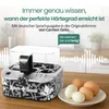 An egg cooker is shown on a wooden countertop with eggs in the foreground. The background is a blurred kitchen scene. Produktbild-Vorschau