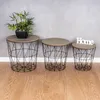 Three wire basket side tables with wooden tops are arranged on a wooden floor against a gray wall. A small potted plant sits on the largest table, and the word "Home" is on the smallest table. Produktbild-Vorschau
