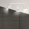 A white track light fixture with three adjustable spotlights, each with a gold accent, is mounted on a white ceiling above a dark wood-paneled wall. Podgląd zdjęcia produktu