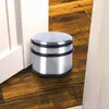 A stainless steel door stopper with a rubber ring sits on a wooden floor next to a white door. Náhled obrázku produktu