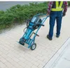 A person in work attire is pulling a heavy-duty demolition hammer on a cart along a paved walkway next to a green hedge. Anteprima dell’immagine prodotto