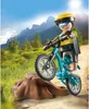 A Playmobil mountain biker figure on a bike is positioned on a dirt path next to a tree stump, with a mountainous landscape in the background. Produktbild-Vorschau