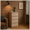 A beige chest of drawers with a clock and plant on top, placed next to a brown sofa against a brick wall. Produktbild-Vorschau