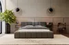A modern bedroom with a dark gray upholstered bed as the focal point, decorated with plants, lamps, and a chair. Produktbild-Vorschau