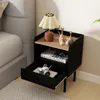 A black nightstand with a drawer and an open shelf, next to a bed. A lamp, decorative cats, and a book are on the nightstand. A camera is in the open drawer. Náhled obrázku produktu