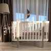A baby crib is set up in a room with sheer curtains and a floor lamp. Produktbild-Vorschau
