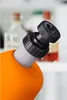 A close-up view of the eyepiece of an orange microscope, with blurred laboratory bottles in the background. Produktbild-Vorschau