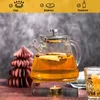 A glass teapot filled with tea is heated by a tealight on a metal stand, with a decorative paper tree and a glass of tea in the background. Produktbild-Vorschau