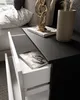 A close-up view of a modern black and white nightstand with an open drawer, revealing a book inside. A decorative sculpture and a vase with dried plants are on top. Produktbild-Vorschau