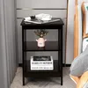 A dark brown wooden nightstand with three shelves is placed next to a bed. The top shelf holds a rolled-up magazine and a white coffee cup. The middle shelf has a small pink vase with flowers. The bottom shelf displays two books, one with 'MARILYN MONROE'  Náhľad obrázku produktu