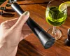 A hand holding a black muddler over a wooden table with a green drink and bar tools. Voir les images du produit
