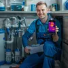 A plumber in blue overalls is kneeling in a room with pipes and water tanks, holding up a smartphone displaying a thermal image. Voir les images du produit