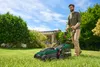 A man is mowing a lawn with a green electric lawnmower on a sunny day. Produktbild-Vorschau