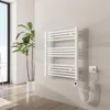 A white electric towel warmer mounted on a light gray tiled wall in a bathroom setting, with a large potted plant to the left. Produktbild-Vorschau