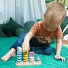 A young child is playing with a wooden stacking toy on a green quilted surface. The background includes green pillows and a white curtain. Produktbild-Vorschau