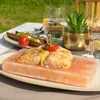 A meal of salmon cooked on a salt block, served outdoors on a table with drinks and a small plant. Produktbild-Vorschau