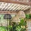 Three woven pendant lamps hang from a wooden ceiling, with lush green plants in the foreground and a stone wall in the background. Náhled obrázku produktu