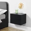 A black floating nightstand with two drawers is mounted on a white wall next to a bed. A glass of water and a wooden clock are on top of the nightstand. Anteprima dell’immagine prodotto