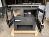 A black wood-burning stove is displayed in a warehouse or workshop setting, with its doors open. Produktbild-Vorschau