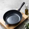 A frying pan with a granite-like finish is placed on a wooden board, with a small bottle of oil and some greenery in the background. Produktbild-Vorschau