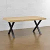 A wooden table with black X-shaped legs is shown against a white brick wall and a wooden floor. Náhled obrázku produktu