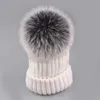 Weiße Strickmütze mit großem schwarz-weißen Kunstfell-Pompon oben, vor grauem Hintergrund Anteprima dell’immagine prodotto