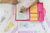 A child's hands are shown using a fashion design kit, with drawings of fashion figures scattered around. Anteprima dell’immagine prodotto