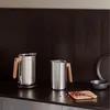 Two stainless steel thermoses with wooden handles on a dark countertop, with some tea leaves scattered on a small wooden board. Voir les images du produit