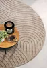 A round, beige rug with a geometric pattern is shown in a living room setting, with a small wooden coffee table holding a candle and a cup of coffee. Anteprima dell’immagine prodotto