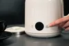 A finger pressing the power button on a white electric kettle, with a black teacup and saucer in the foreground. Podgląd zdjęcia produktu