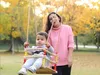 A woman stands behind a child sitting on a wooden swing in a park. Produktbild-Vorschau