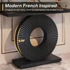 A black decorative circular object with a fluted design sits on a black rectangular base, placed on a marble-like surface. The object is positioned in front of a framed background element. Text overlays describe the product as 'Modern French Inspired' and  Náhľad obrázku produktu