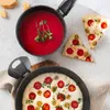 A top-down view of a black aluminum saucepan filled with red tomato soup, garnished with fresh herbs. Next to it are two slices of focaccia pizza topped with cherry tomatoes and olives. Another black frying pan with a similar focaccia pizza is visible in t Produktbild-Vorschau