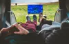 Two people relaxing in the back of a vehicle, watching a TV screen set up outside. Produktbild-Vorschau