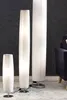 Three white floor lamps of varying heights are arranged in a room with a dark gray wall and a patterned rug. Produktbild-Vorschau