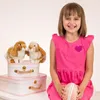 A young girl in a pink dress sits with two stuffed bunnies on top of two white suitcases. She is holding a pink and white patterned egg-shaped object. Anteprima dell’immagine prodotto