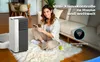 A woman is sitting on a fluffy rug in a living room, holding a smartphone and looking at the camera. Next to her is a portable air conditioner. The background shows a sofa and part of a dining area. Produktbild-Vorschau