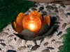A solar-powered garden light shaped like a lotus flower with an orange, cracked glass orb in the center, placed on a bed of white and gray pebbles with some green grass in the background. Produktbild-Vorschau