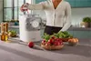 A woman is pouring liquid into a pasta maker with fresh ingredients like radishes, spinach, and eggs on a kitchen counter. Náhľad obrázku produktu
