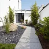 A modern entryway with a stone pathway leading to a dark gray door, flanked by bamboo plants and large rocks. Produktbild-Vorschau