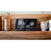 A black digital radio is placed on a wooden shelf next to a brown glass bottle and two mugs. Produktbild-Vorschau