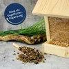 A wooden bird feeder filled with birdseed, with a pile of loose birdseed and a wooden scoop in the foreground, and decorative grass and a piece of driftwood in the background. A circular graphic with text is overlaid on the left side. Produktbild-Vorschau