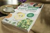 A cookbook titled "Basisch kochen mit dem Thermomix®" is displayed on a textured brown cloth, with a cup of coffee and a small plant in the background. Produktbild-Vorschau