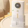 A white air purifier with a gold accent ring around a digital display sits on a light-colored table, with decorative green leaves and sparkling particles floating around it. Produktbild-Vorschau