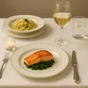 A formal dinner setting with a plate of salmon and spinach, a side of pasta, a glass of white wine, and a lit candle. Produktbild-Vorschau